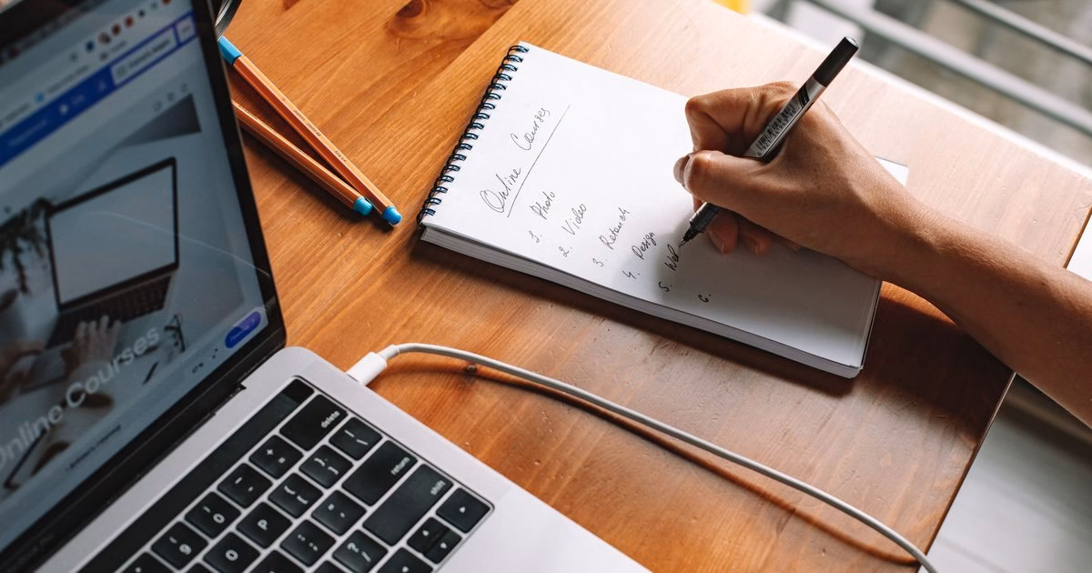 Persona anotando una lista de cursos online en un cuaderno, junto a un notebook que muestra una plataforma de e-learning